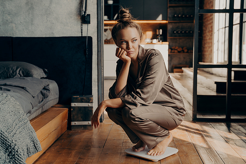 Young dissatisfied unhappy woman crouched over electronic scales in morning after waking up in modern bedroom background, displeased with her weight goals. Dieting concept