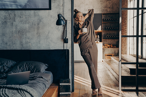 Full length of attractive relaxed caucasian young female in satin pajama with cup of coffee in her hand standing stretching in bedroom doorway, enjoying early morning wake up and start of work week