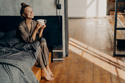 Starting new beautiful day. Cute pretty woman with cup of morning coffee chilling while sitting on bed, relaxing in brown satin pajama in modern apartment. Happy time at home