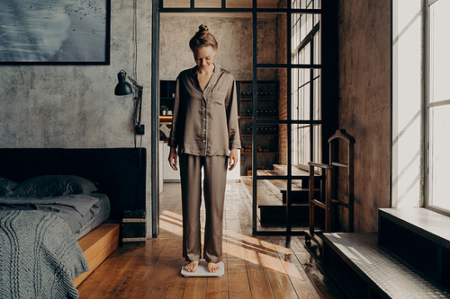 Young lovely female in comfy satin pajama standing on electronic scales in bedroom after waking up in early morning before taking refreshing shower. Weightloss and healthy lifestyle concept