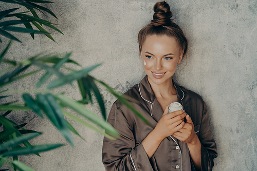 Image of caucasian cheerful woman in silk brown pajama smiling while applying face cream isolated over concrete wall background. Daily routine procedure for rejuvenation and care of skin