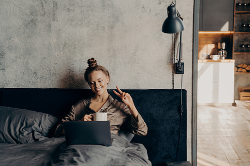 Lovely joyful young girl lying in bed after waking up in early morning, having cup of hot coffee while having video call with friends online on laptop, smiling and showing V sign with her fingers