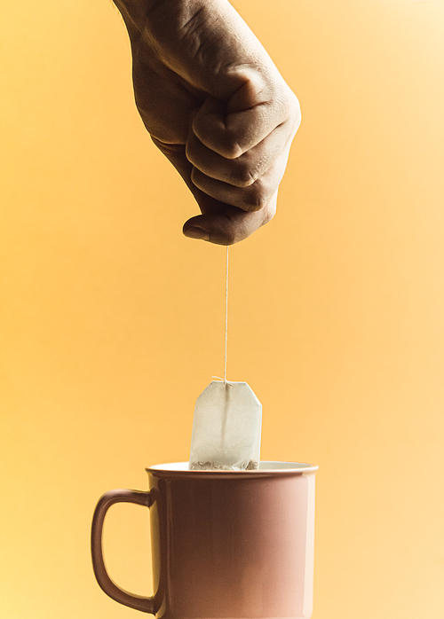 A super minimalistic shot of a hand holding a bag of fast tea over a pink cup over a pastel yellow background