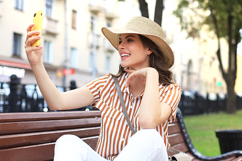 Portrait of pretty young woman making selfie by the phone sitting on bench in the city street