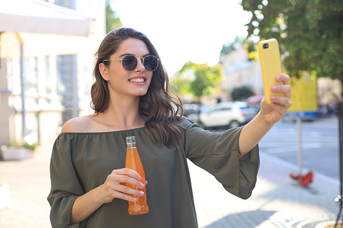 Smiling beautiful brunette woman walking outdoors by street, take selfie by mobile phone, drinking juice