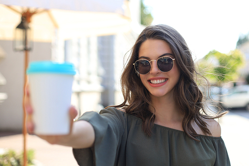 Photo of smiling cute woman in green dress holding paper cup, walking outdoors by street