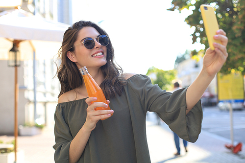 Smiling beautiful brunette woman walking outdoors by street, take selfie by mobile phone, drinking juice