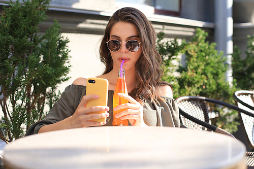Smiling beautiful brunette woman sitting in street summer cafe, take selfie by mobile phone, drinking juice
