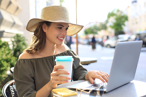 Happy nice woman working on laptop in street cafe, holding paper cup