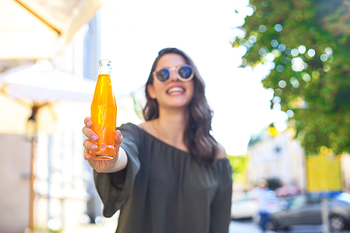 Attractive young woman in sunglasses holds a bottle with juice, looking at camera, outdoors