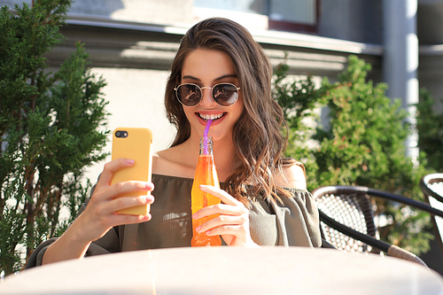 Smiling beautiful brunette woman sitting in street summer cafe, take selfie by mobile phone, drinking juice