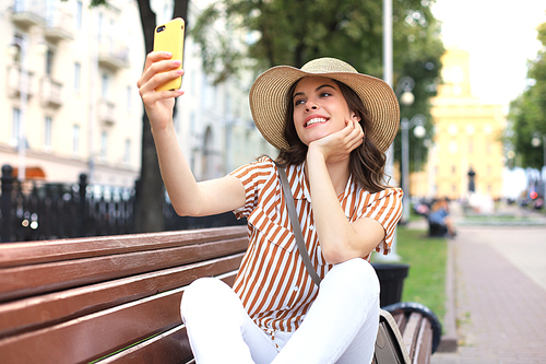 Portrait of pretty young woman making selfie by the phone sitting on bench in the city street