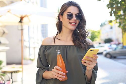 Attractive young woman in sunglasses holds a bottle with juice, using smartphone, outdoors