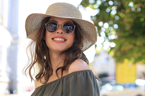 Smiling pretty girl wearing sunglasses and summer hat and looking at camera while walking in city street