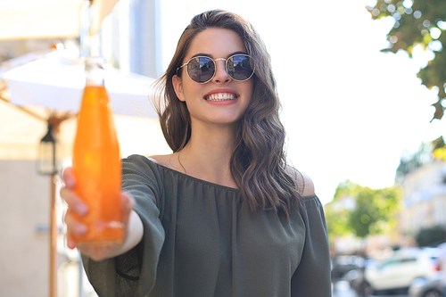 Attractive young woman in sunglasses holds a bottle with juice, looking at camera, outdoors