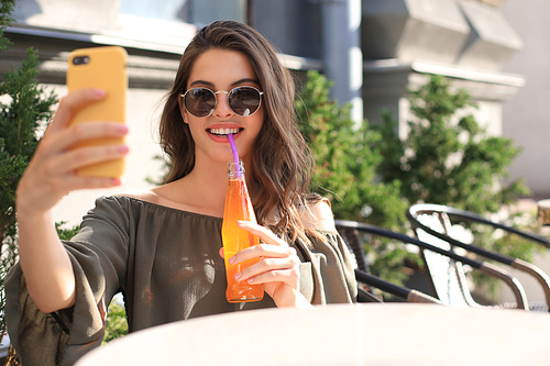 Smiling beautiful brunette woman sitting in street summer cafe, take selfie by mobile phone, drinking juice