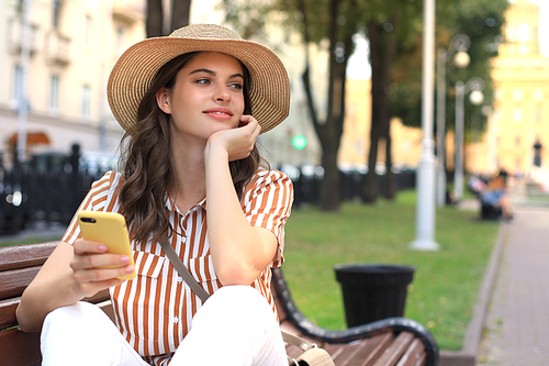 Beautiful happy young woman relaxing on the bench at the city park, holding mobile phone in hands