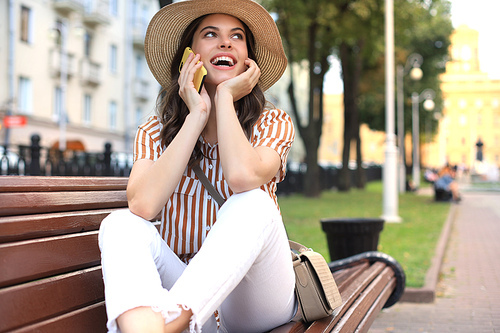 Attractive laughing woman talking on cellphone while sitting on bench outdoors.