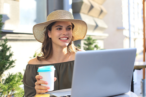 Happy nice woman working on laptop in street cafe, holding paper cup