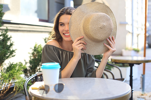 Portrait of a girl hiding her face behind a straw hat while sitting in summer street cafe