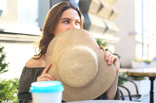 Portrait of a girl hiding her face behind a straw hat while sitting in summer street cafe
