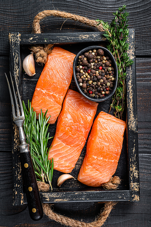 Raw salmon fillet fish steaks in a wooden tray with thyme and rosemary. Black wooden background. Top view.