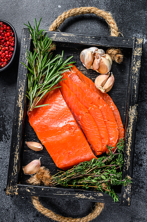 Smoked sliced salmon fillet in a wooden tray with herbs. Black background. Top view.