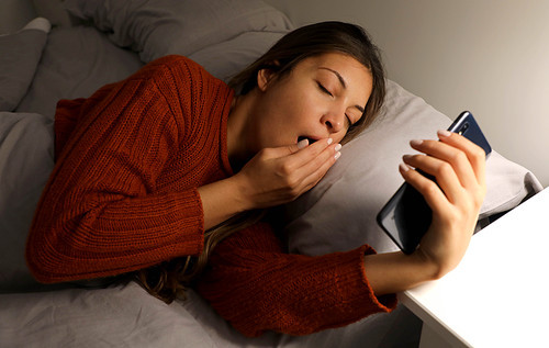 Beautiful young woman lying on bed and yawning looking social media on smart phone. Tired girl watching mobile or listening music and sleepy, female lazy and bored, lifestyle concept.