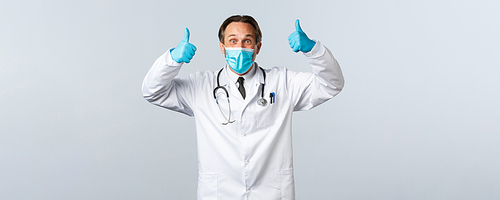Covid-19, preventing virus, healthcare workers and vaccination concept. Enthusiastic happy and cheerful male doctor in medical mask and gloves cheering, show thumbs-up in approval or recommendation.
