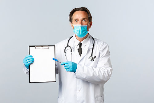 Covid-19, preventing virus, healthcare workers and vaccination concept. Shocked male doctor in medical mask and gloves pointing at clipboard, stare astonished, showing important information.
