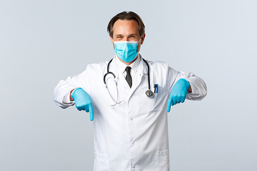 Covid-19, preventing virus, healthcare workers and vaccination concept. Pleasant doctor in medical mask and gloves give advice, recommend promo, pointing fingers down at banner.