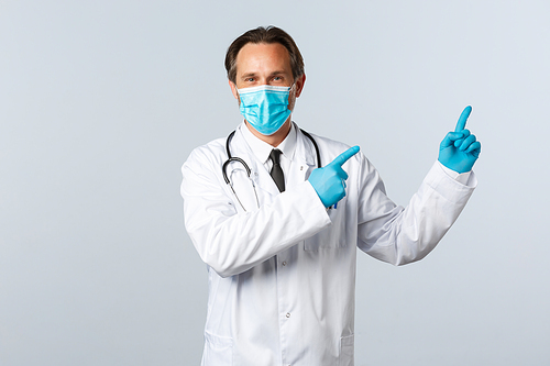 Covid-19, preventing virus, healthcare workers and vaccination concept. Smiling friendly doctor in white coat, medical mask and gloves pointing top right advertisement, white background.