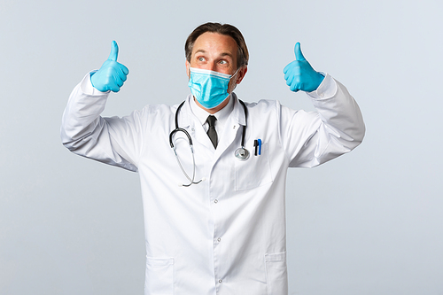 Covid-19, preventing virus, healthcare workers and vaccination concept. Cheerful pleased doctor in medical mask and gloves show thumbs-up and looking left satisfied, white background.