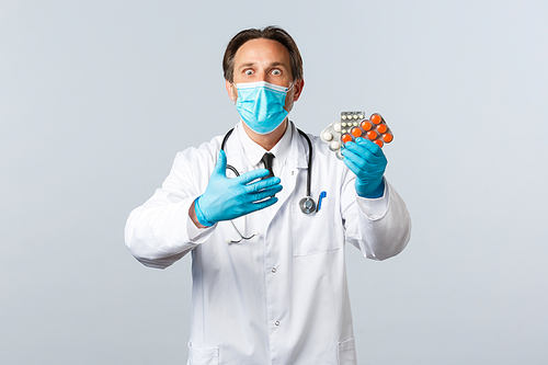 Covid-19, preventing virus, healthcare workers and vaccination concept. Excited and shocked doctor in medical mask and gloves react to new medication, showing pills, white background.
