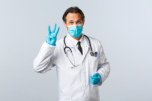 Covid-19, preventing virus, healthcare workers and vaccination concept. Serious caring doctor in medical mask and gloves explain rules during social distancing, show three fingers, white background.