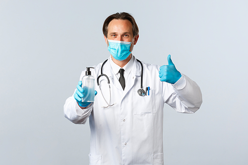 Covid-19, preventing virus, healthcare workers and vaccination concept. Satisfied doctor in medical mask and gloves thumb-up in approval, recommend hand sanitizer, white background.