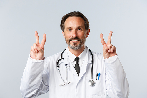 Covid-19, preventing virus, healthcare workers and vaccination concept. Cheerful handsome middle-aged doctor in white coat showing peace signs or v-sign and smiling, stay positive.