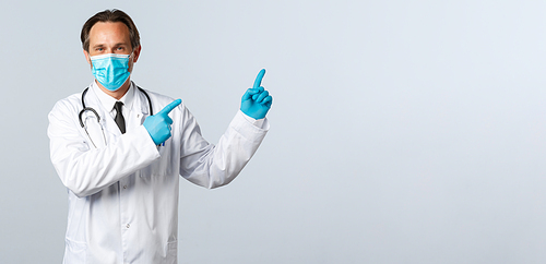 Covid-19, preventing virus, healthcare workers and vaccination concept. Smiling friendly doctor in white coat, medical mask and gloves pointing top right advertisement, white background.