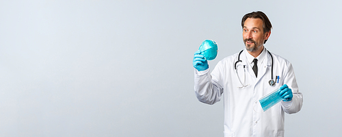 Covid-19, preventing virus, healthcare workers and vaccination concept. Enthusiastic middle-aged doctor in white coat and gloves, showing two PPE, looking at medical respirator pleased, hold mask.