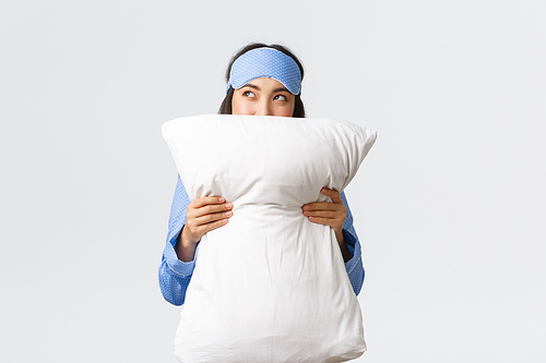 Dreamy kawaii asian girl in sleeping mask and blue pajamas hugging pillow and looking cunning at upper left corner, imaging something, having idea in comment bubble, standing white background.