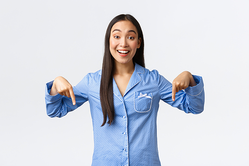 Cheerful smiling asian girl react to wonderful news, pointing fingers down as standing in blue pajamas, showing girlfriends cool thing at sleepover party, making announcement, white background.