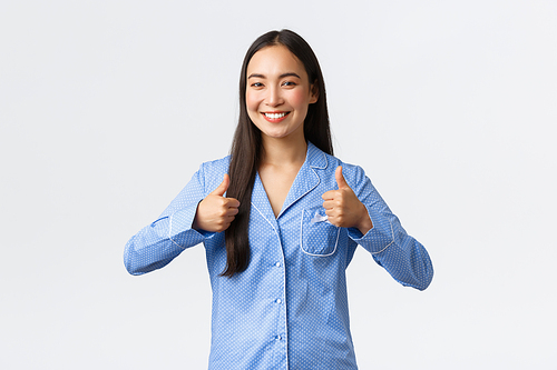 Upbeat happy smiling asian girl in blue pajamas looking satisfied, showing thumbs-up in approval, saying well done or good job, standing pleased over white background.