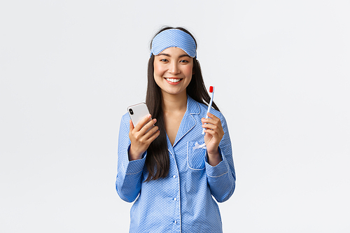 Hygiene, lifestyle and people at home concept. Smiling cute asian girl in blue pajamas and sleeping mask, brushing teeth before bed and using smartphone, showing white teeth, white background.