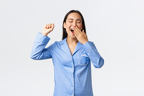 Smiling cheerful asian woman in blue jammies, yawning cover opened mouth with palm and stretching hands up after waking-up in morning. Girl ready start new day after good nap, white background.