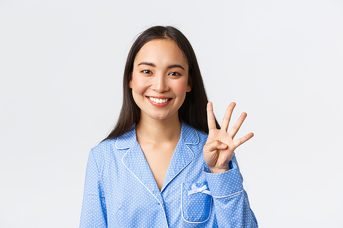 Close-up shot of happy attractive asian woman in blue pyjama showing four fingers and smiling white teeth, explain main rules or making order, standing white background delighted.