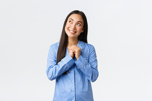 Dreamy cute asian girl in blue pajamas smiling cheerful and gazing upper left corner, picturing nice moment, imaging thing before sleep, looking pleased and standing over white background.