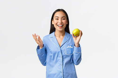 Morning, active and healthy lifestyle and home concept. Excited asian girl in blue pajamas looking happy, smiling as showing apple, eating fruits for breakfast, white background.