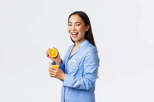 Morning, active and healthy lifestyle and home concept. Excited happy asian girl in pajamas smiling at camera while squeezing orange in glass, drinking orange juice over white background.