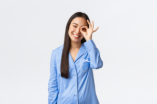 Happy beautiful asian woman in blue pajamas with perfect clean skin and whtie teeth, showing okay gesture over eye with satisfied expression, guarantee quality, recommend product, being pleased.