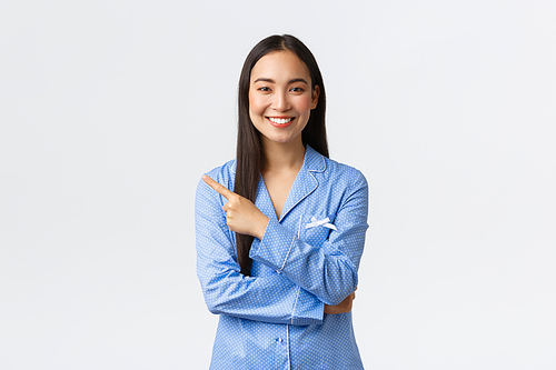 Beautiful young asian girl in blue pajamas pointing finger left at promo banner. Woman in jammies showing way or announce discount at store with women care products, white background.